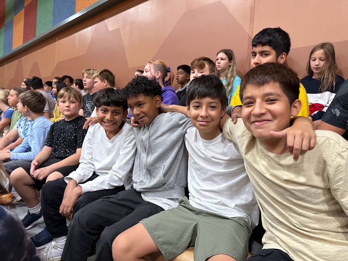 Many students sit in the bleachers for an assembly. Four boys link arms over shoulders to pose for the picture.
