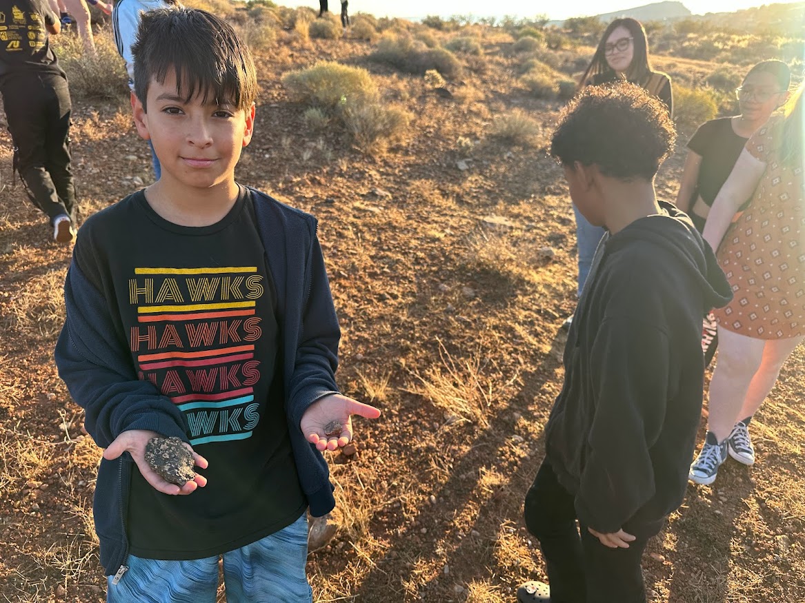 One seventh grade male student shows off his two rock finds. In the background are more students searching the desert.