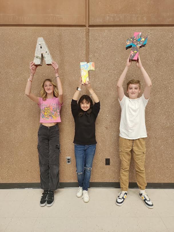 Three students hold up their 3D letter creations in the commons area. The three letters spell ART.