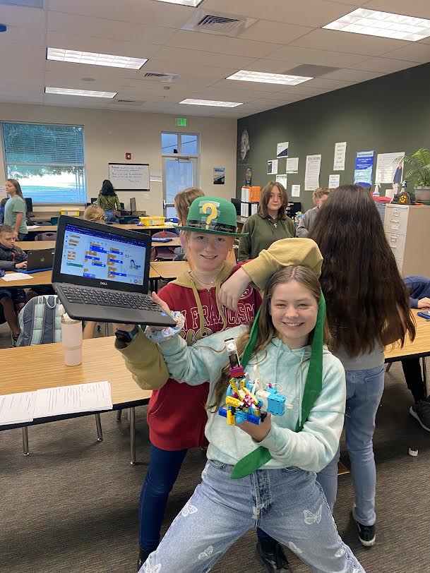 Two 6th grade female students pose for the camera showing off their completed Lego robot and coding. In the background of Mr. White's Creative Coding classroom you can see several other students working on their Lego robots.