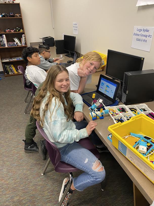 Two 6th grade female students smile for the camera during Creative Coding in Mr. White's classroom. Two male students concentrate on their own robot in the background.