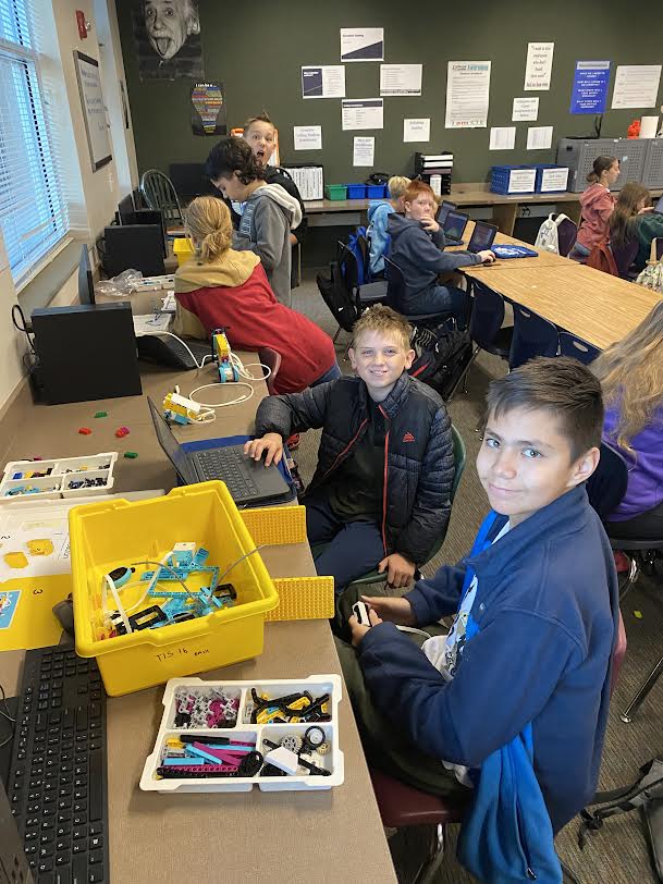 Two 6th grade male students smile for the camera during Creative Coding in Mr. White's classroom. All 10 students are working on their Lego robots.